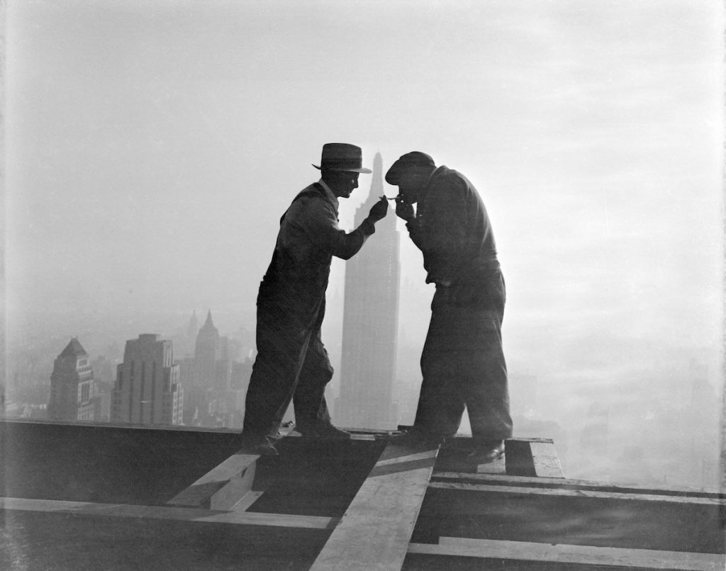 2 decembrie 1932, doi muncitori care lucrau la clădirea RCA își aprind țigările. În fundal, silueta Empire State Building, ce arată înălțimea la care se aflau cei doi. | Imagine originală, Credit: Bettmann Archive, Sursa: History Collection