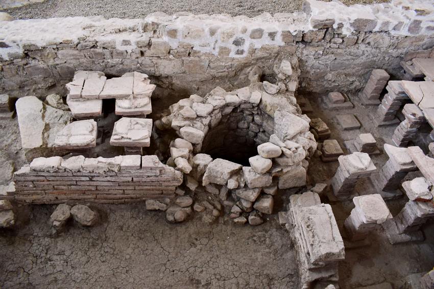 În Muzeul Principia: În dreapta imaginii: Hypocaust, încălzire prin podea. În zidul exterior, era o vatră de unde fumul și căldura circula pe dedesubtul podelei și o încălzea.  În mijloc: Fântâna e medievală, datată prin secolele XI-XVV – un mic exemplu de refolosire a pietrei romane. „S-a construit rudimentar, n-au prea evoluat oamenii timp de o mie de ani, au pierdut rețeta betonului de la romani, pietrele la fântână au fost lipite cu pământ”, explică Ciprian. | Credit foto: Mira Kaliani